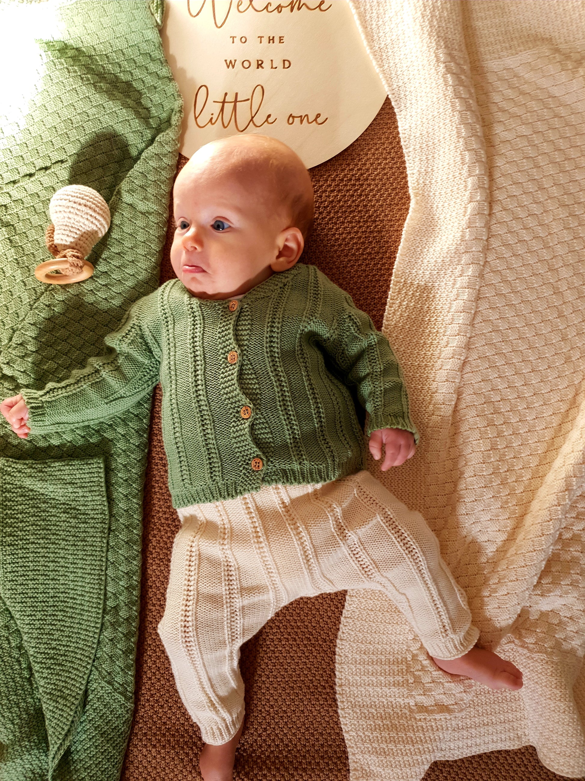 A baby in a green knitted sweater and cream pants.