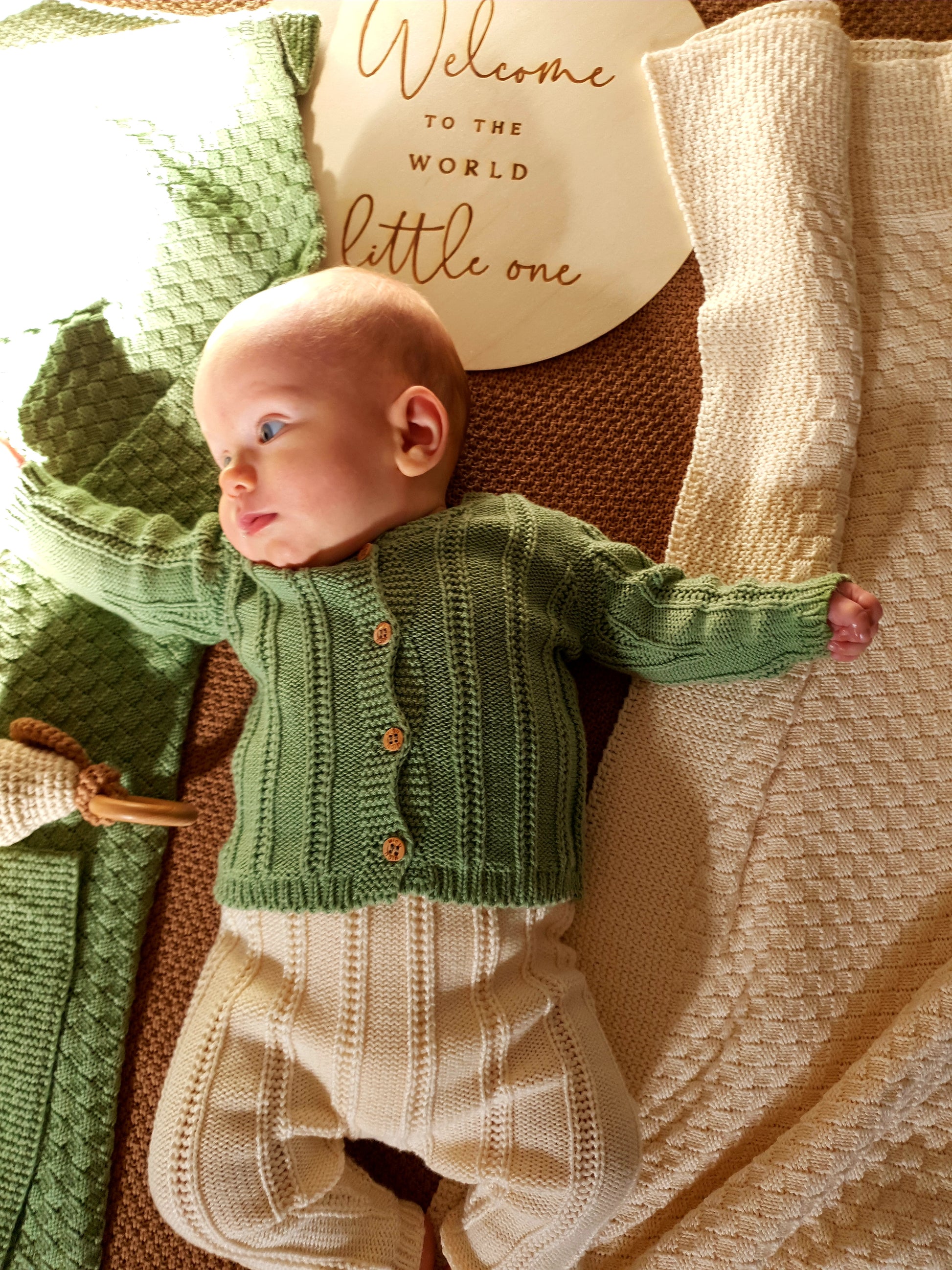 A baby in a green sweater and cream pants lies on a blanket.