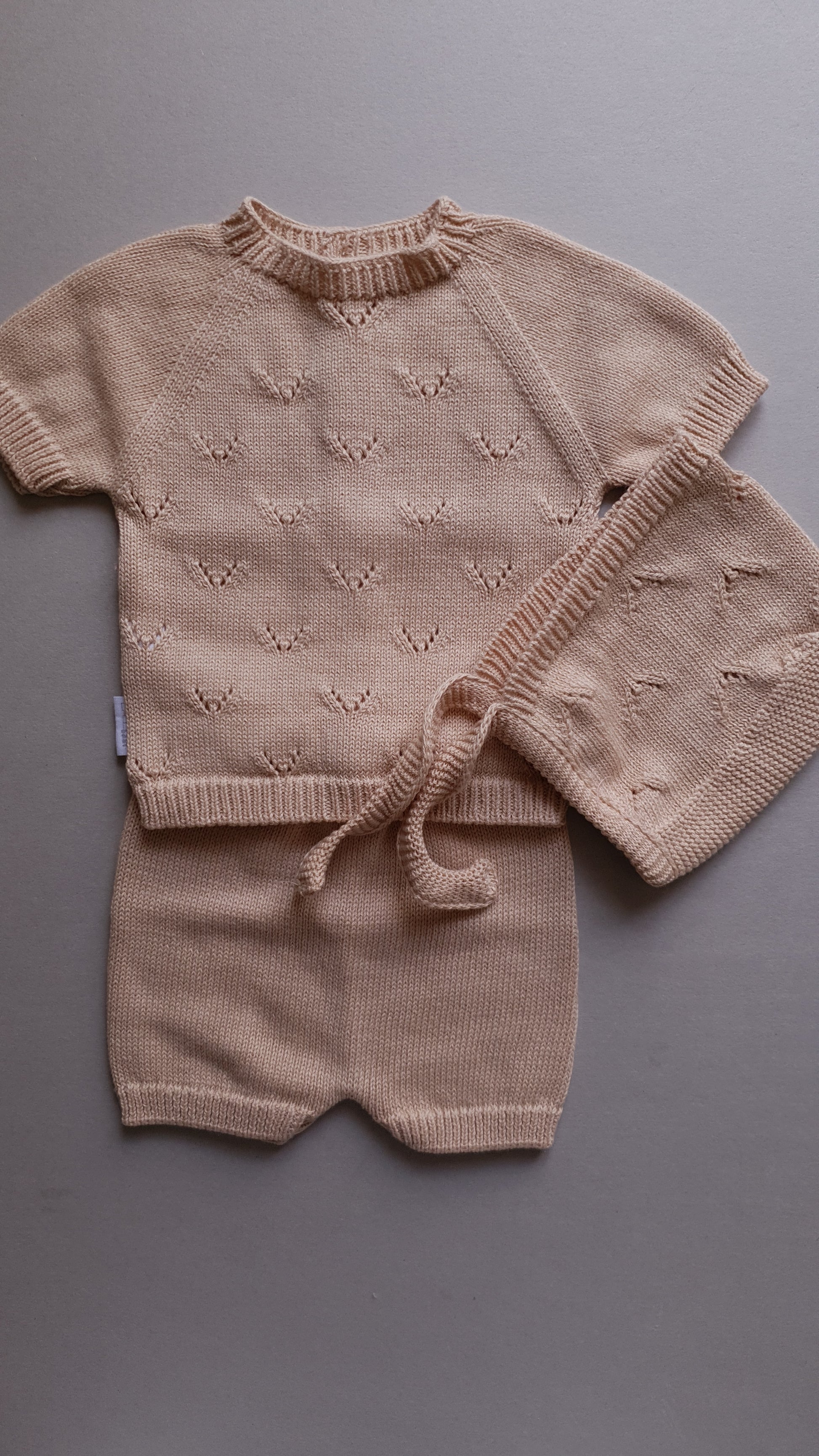 Beige baby outfit with short sleeves, a bonnet, and shorts.