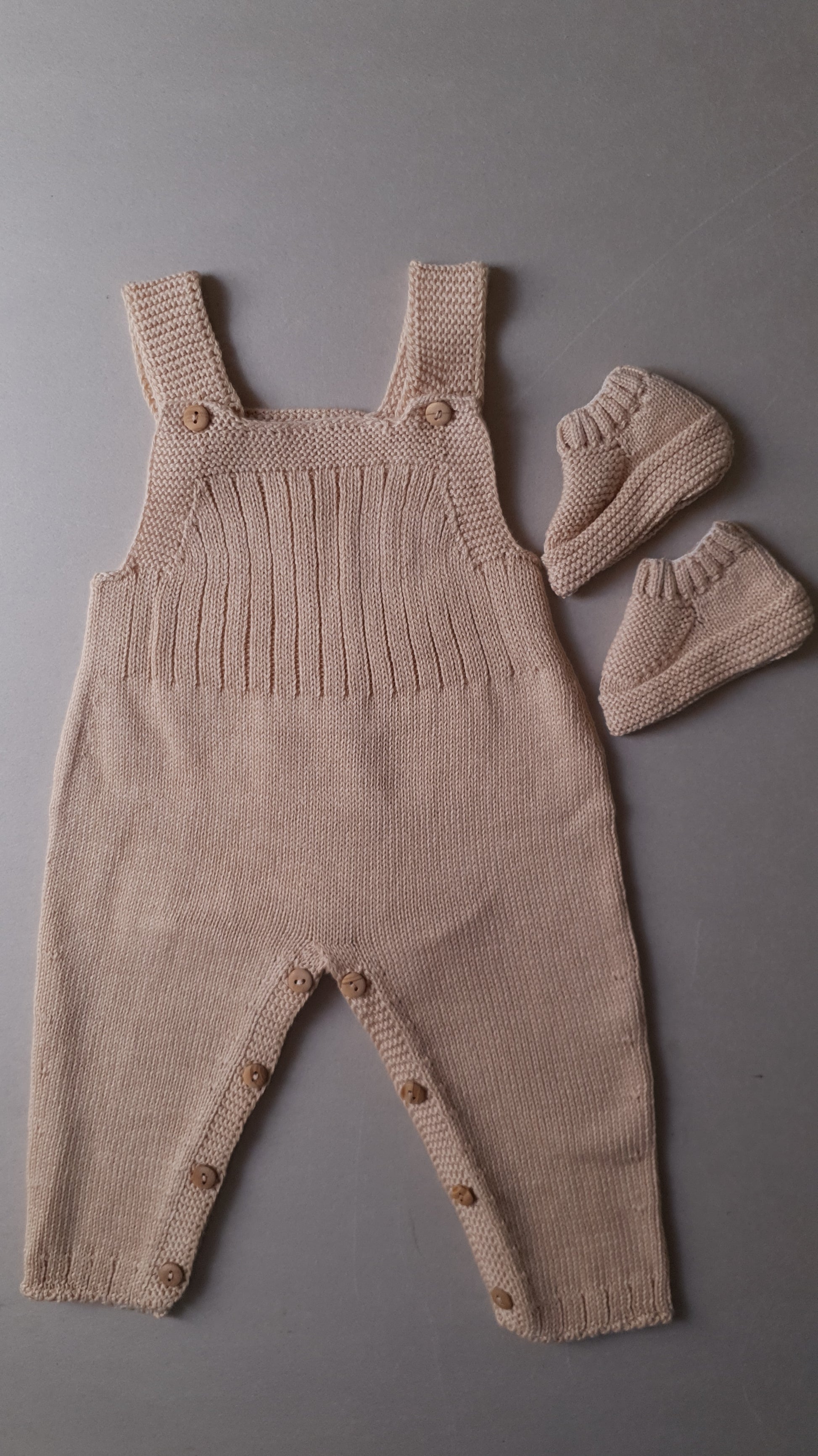 Beige knitted baby outfit with shoulder straps and buttons.