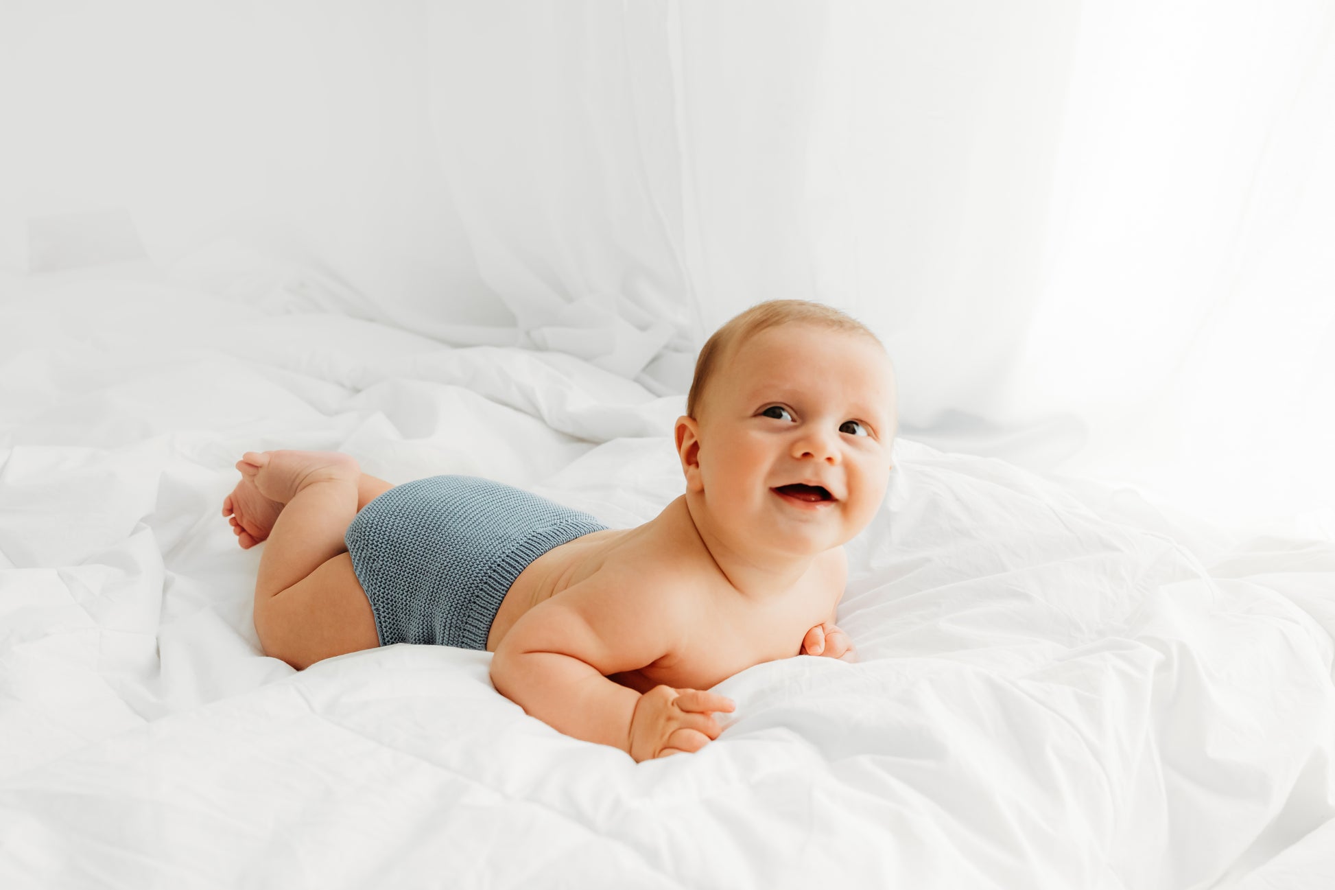 A smiling baby wearing a blue diaper lies on a fluffy bed.