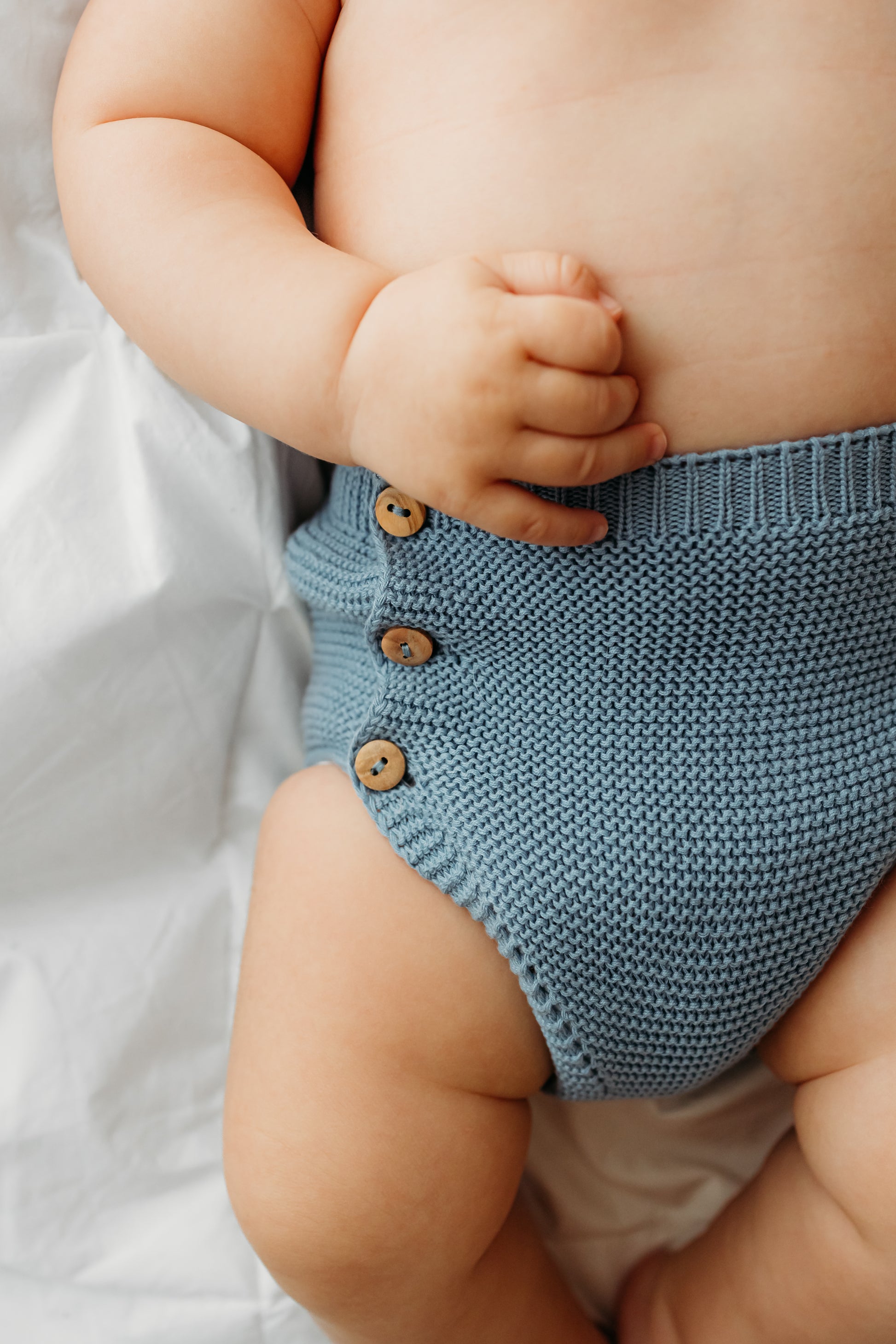 A baby wearing knitted blue shorts with wooden buttons.
