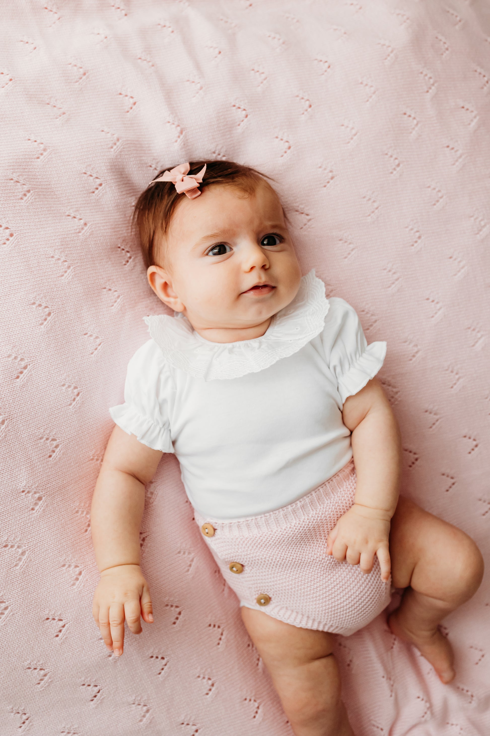 A baby wearing a white ruffled top and pink knit shorts.