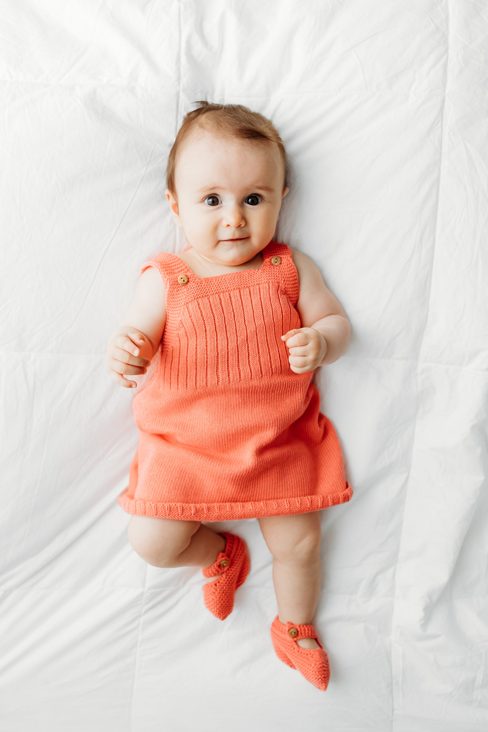 A baby in a coral knitted dress lies on a white blanket.