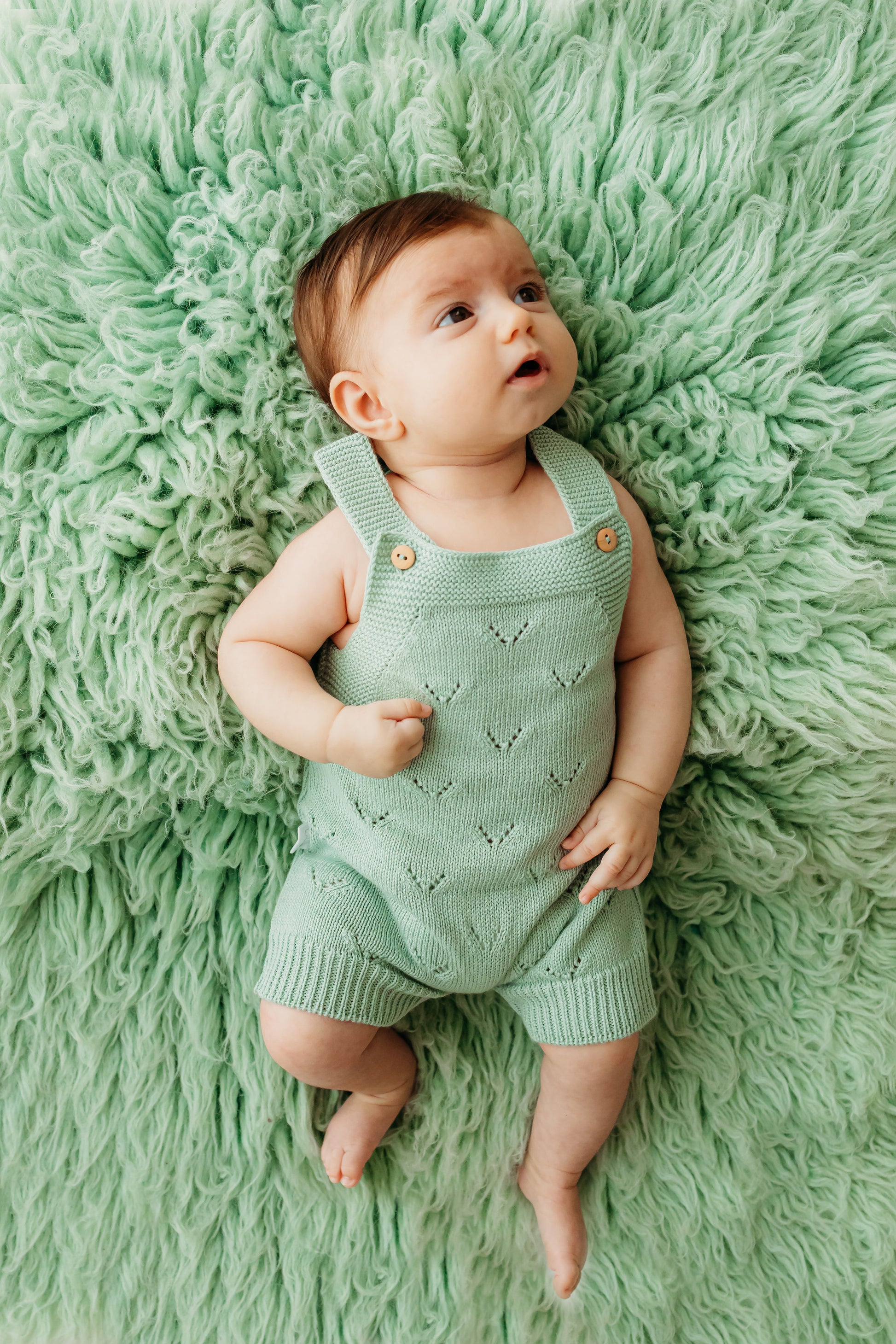 A baby in a mint green knitted romper with wooden buttons.