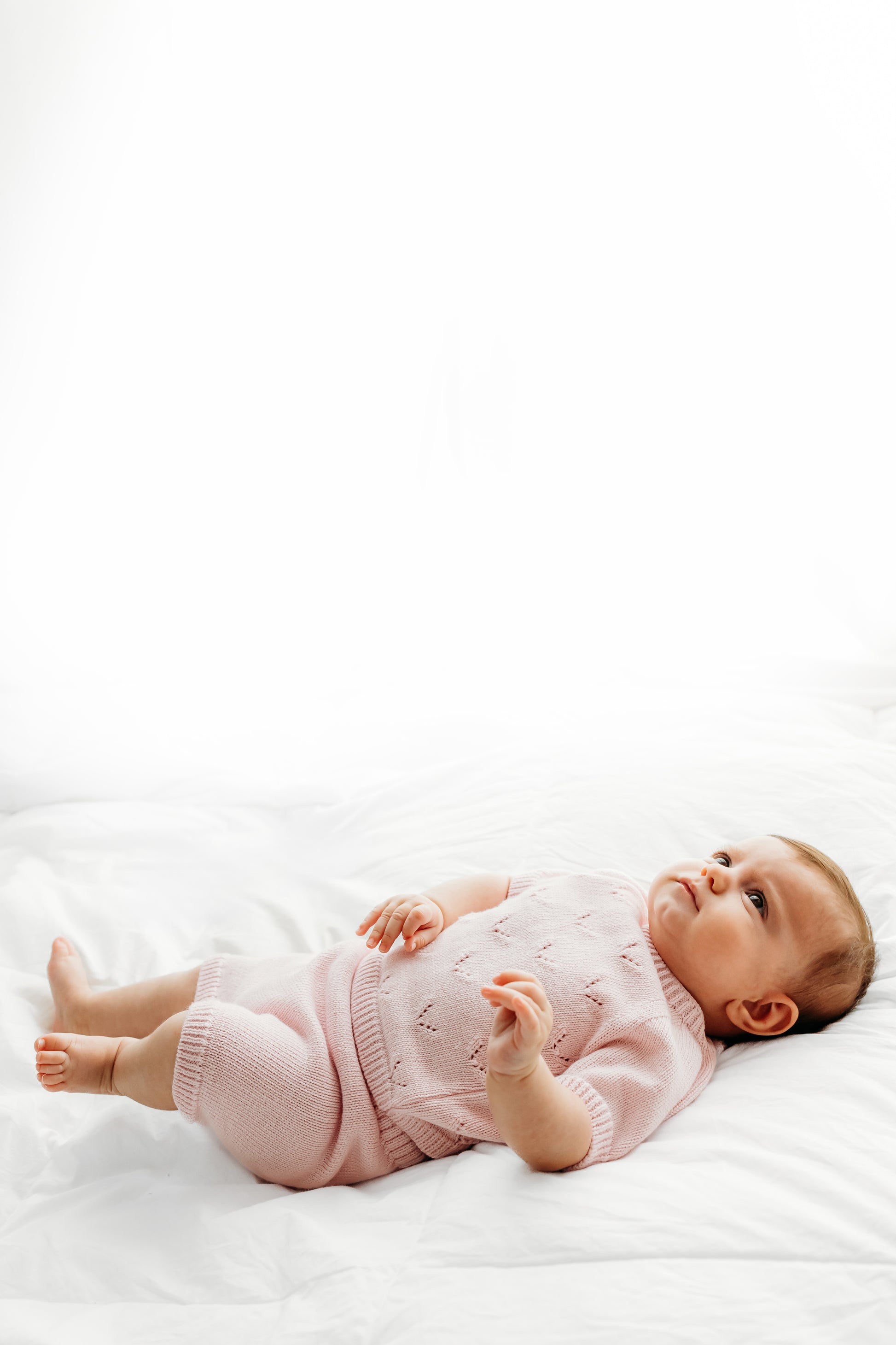 A baby, dressed in a pink outfit, lying on a white blanket.