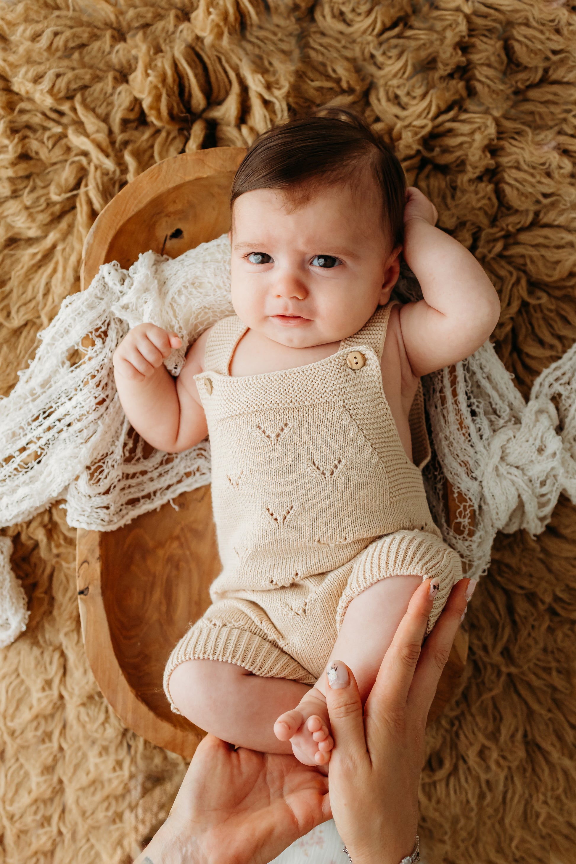 A baby wearing a beige romper lies on a fluffy, tan blanket.