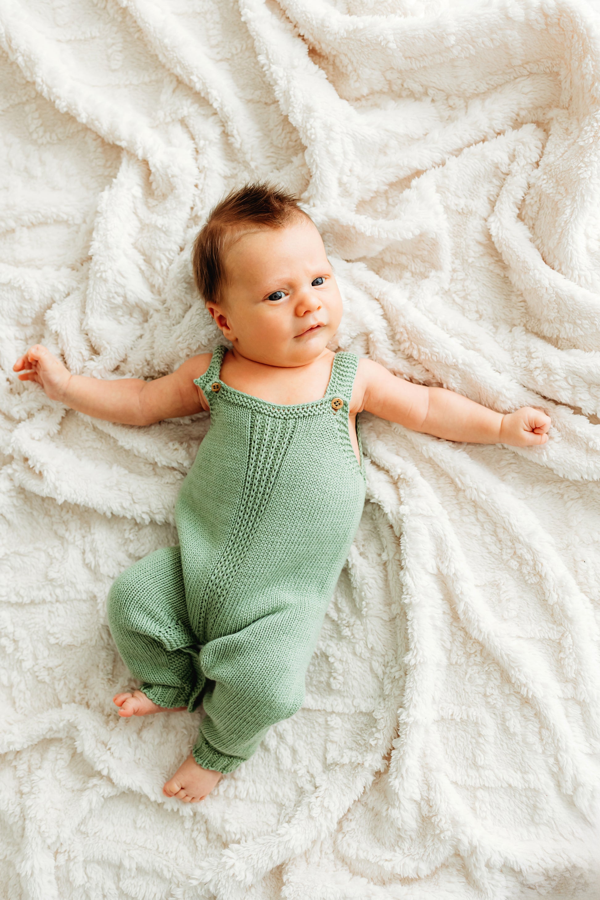 A baby on a white blanket, wearing a green knitted romper.