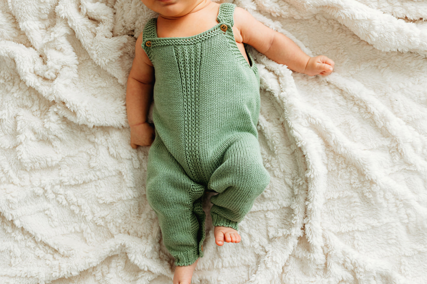 A baby in a green knitted outfit lies on a cream blanket.