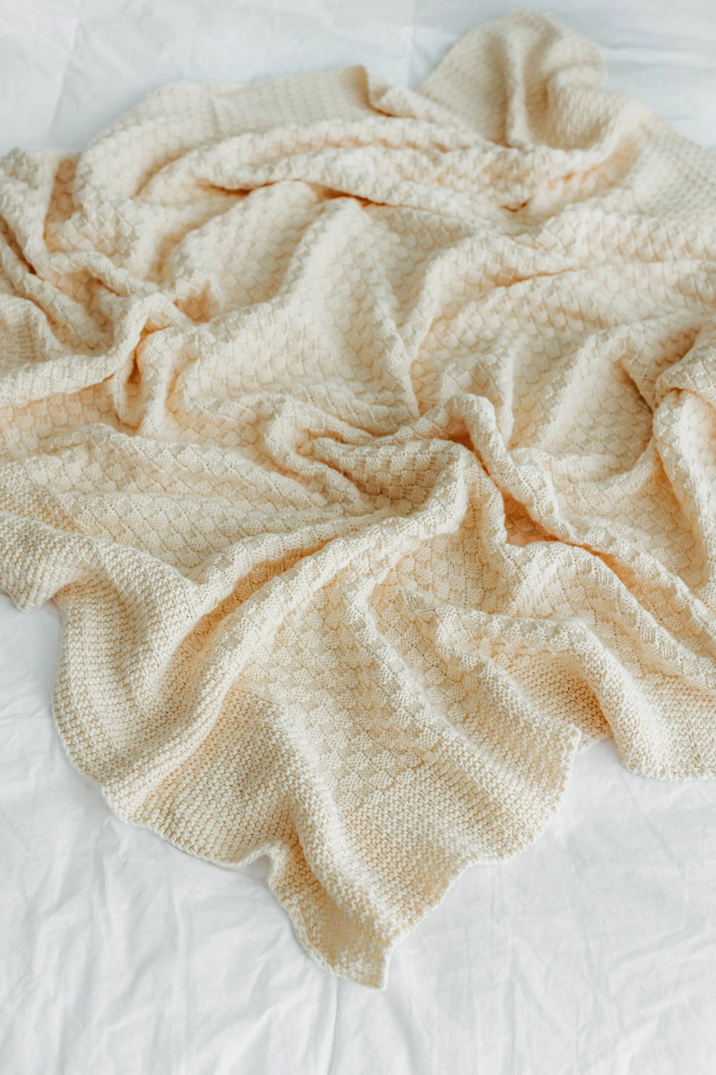 Cream-colored textured blanket crumpled on a white bedsheet.