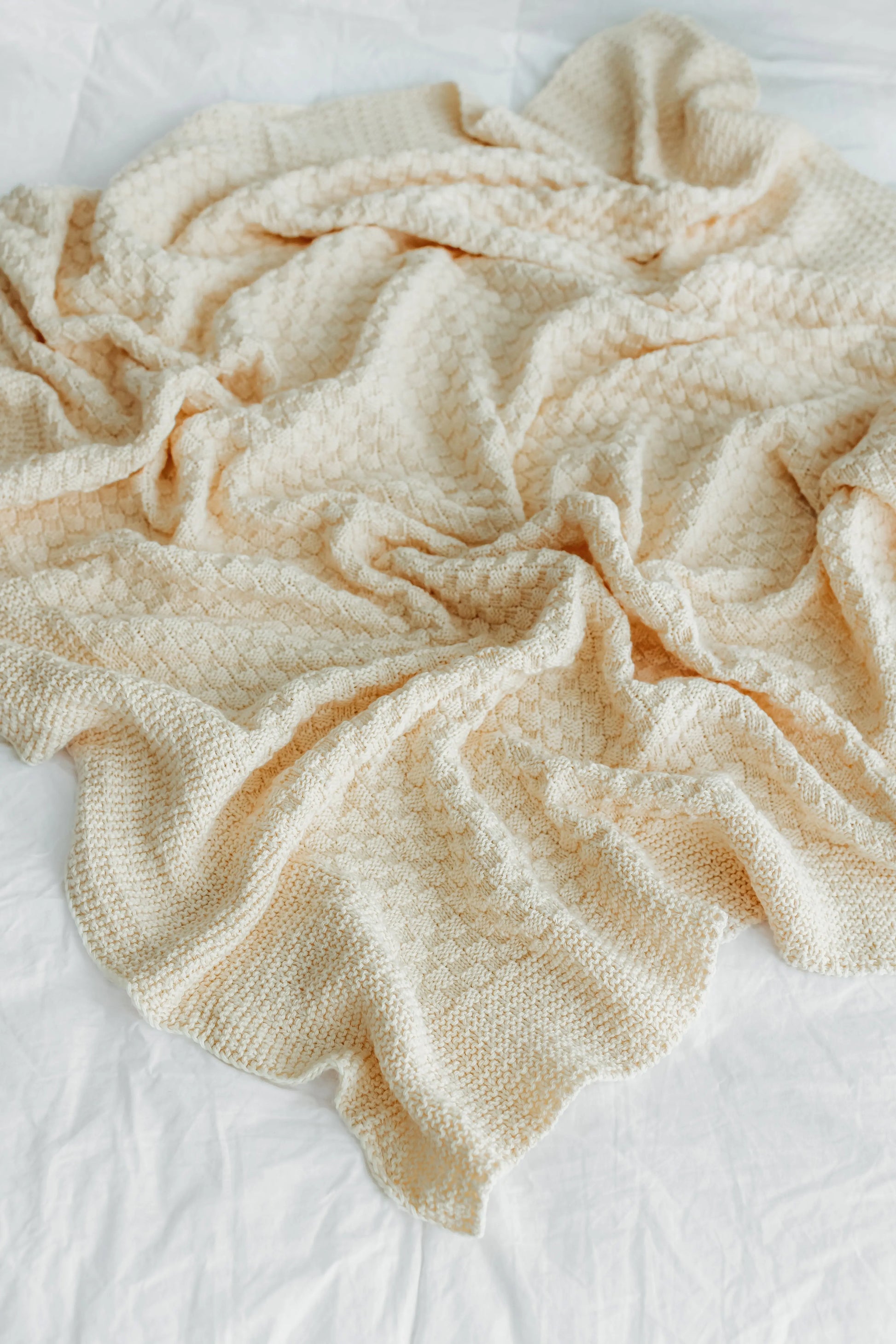 Cream-colored textured blanket crumpled on a white bedsheet.