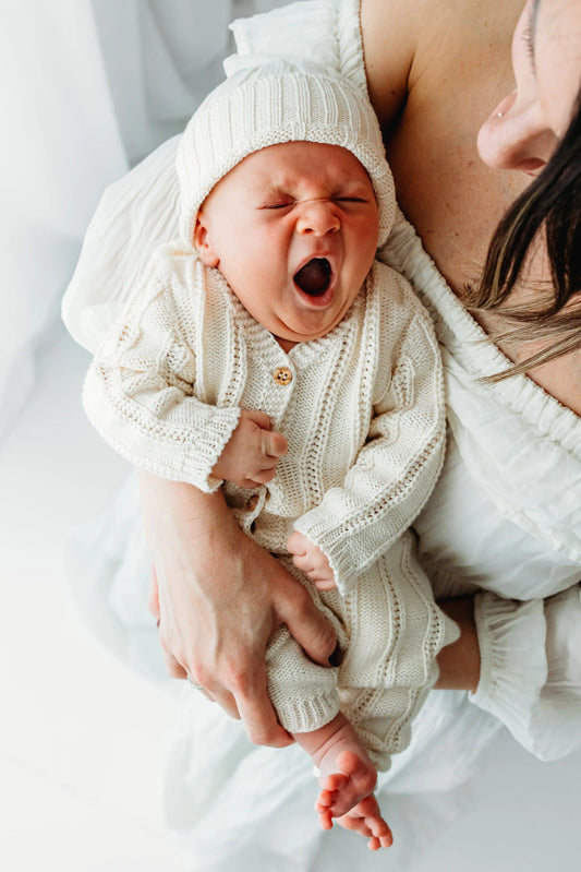 A baby wearing a knit outfit and hat is yawning.