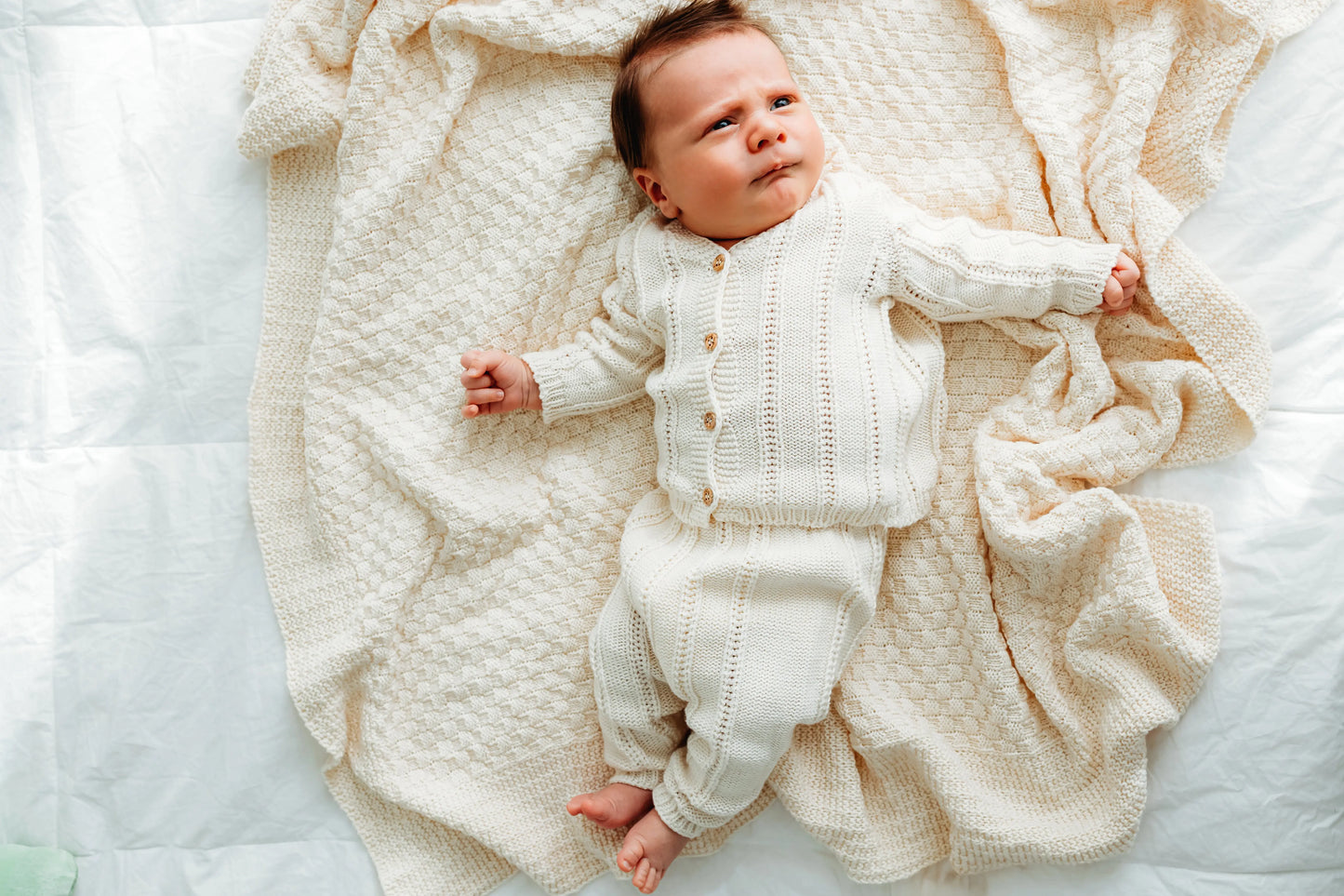 A baby in a cream outfit lies on a matching cream blanket