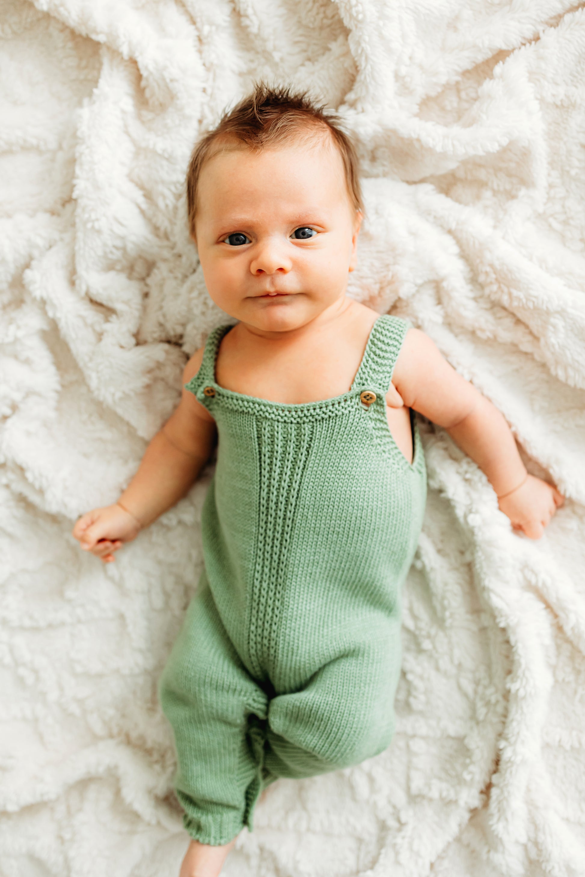 A baby in a green knitted romper lies on a white blanket.