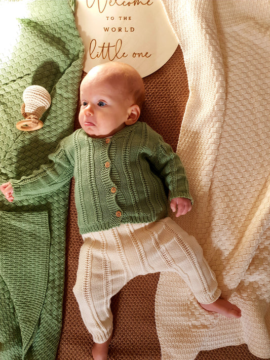 A baby in a green knitted sweater and cream pants.