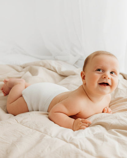 A smiling baby wearing a blue diaper lies on a fluffy bed.