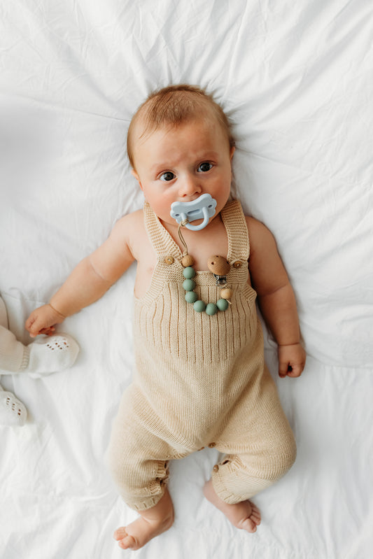 A baby lying on a white blanket wears a beige knit romper.