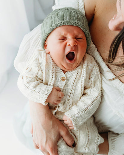 A baby wearing a knit outfit and hat is yawning.