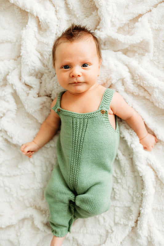 A baby in a green knitted romper lies on a white blanket.
