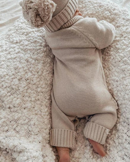 A baby in a beige knit outfit and hat lies face down.